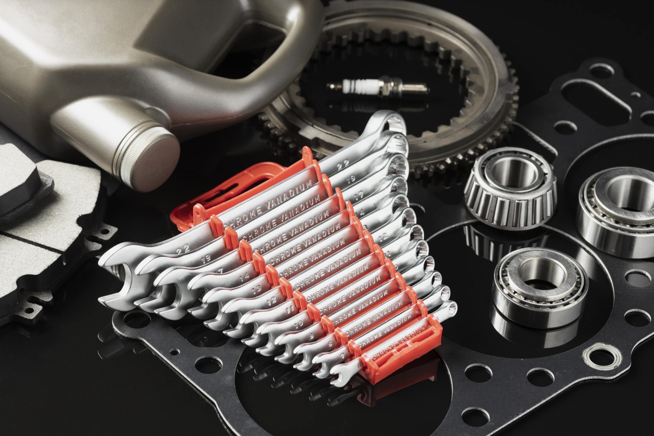 Assorted automotive tools and parts including wrenches, spark plug, bearings, brake pads, and gear components arranged on a black surface.