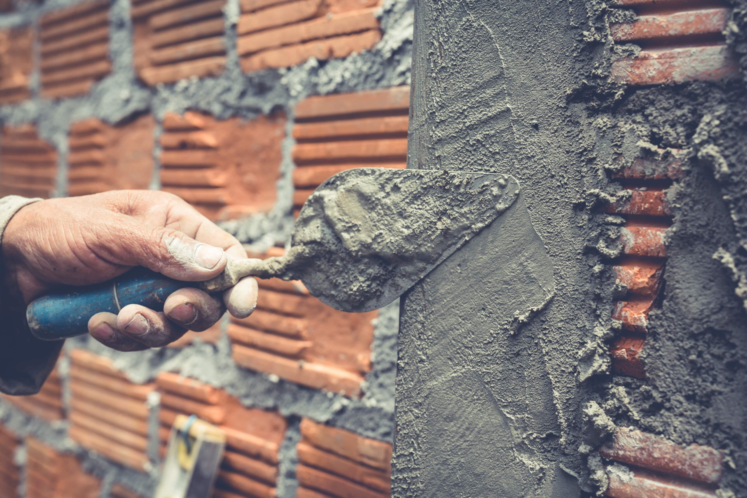 Close-up of a hand applying mortar to a red brick wall using a trowel during masonry work.