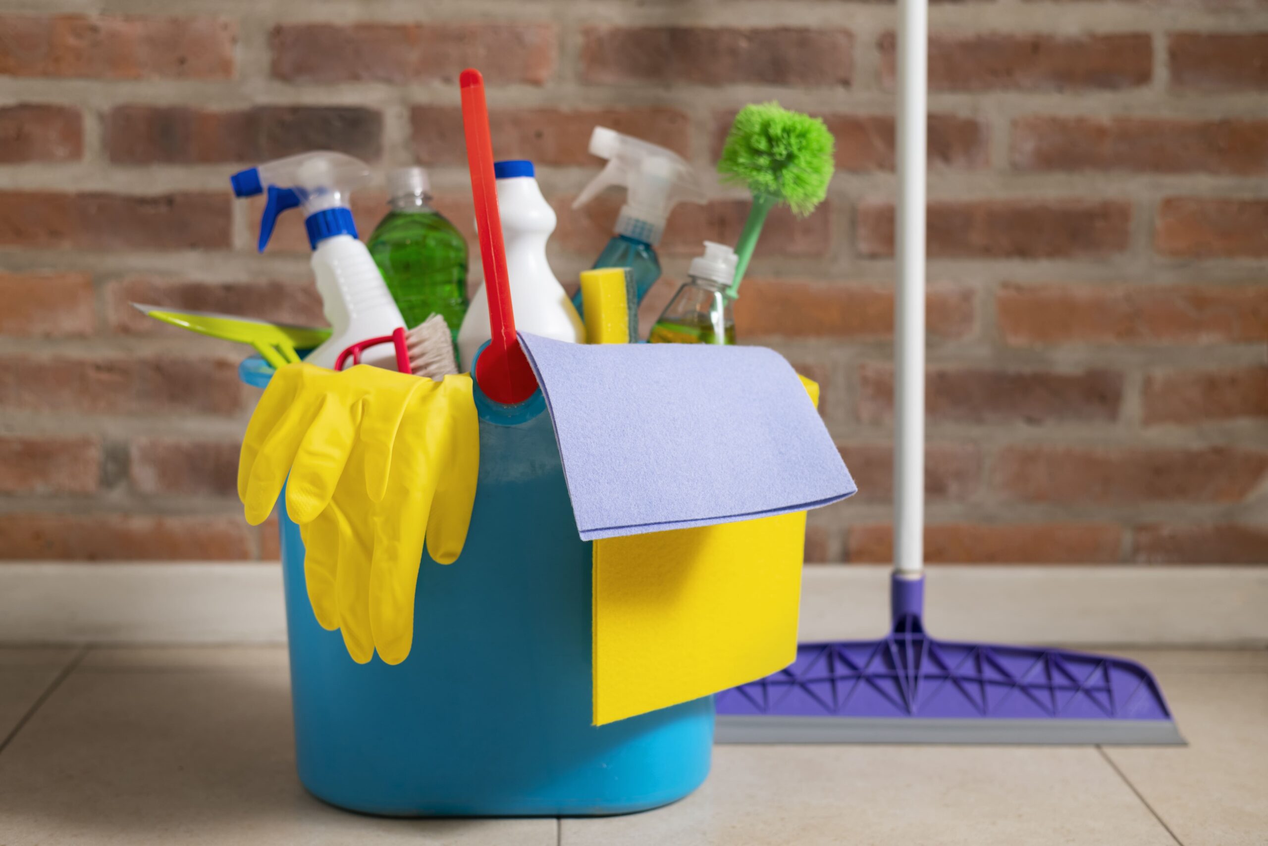 Blue bucket filled with cleaning supplies including gloves, spray bottles, brushes, cloths, and a mop, set against a brick wall and tiled floor.