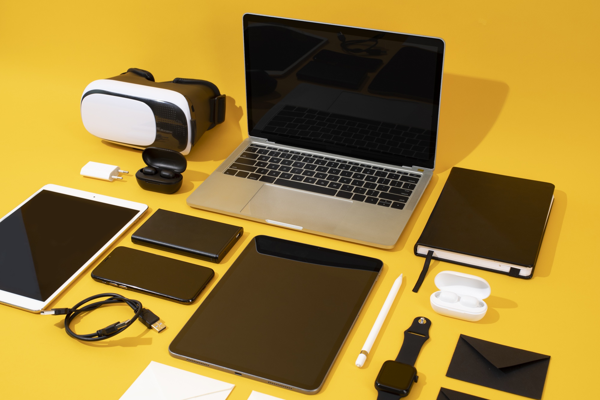 Assorted consumer electronics including a laptop, VR headset, tablet, smartphone, smartwatch, and wireless earbuds arranged on a yellow background