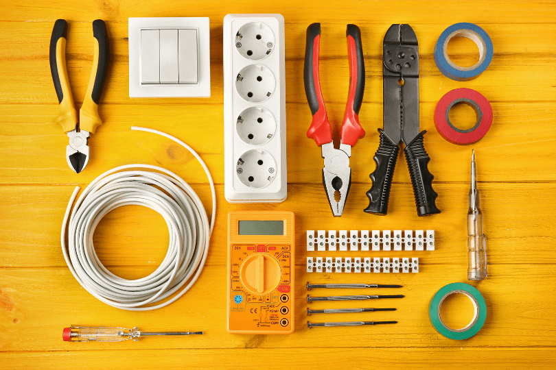 Assorted electrical tools and components including wire cutters, pliers, multimeter, screwdrivers, power strip, light switch, electrical tape rolls, coiled cable, and terminal blocks arranged on a yellow wooden surface