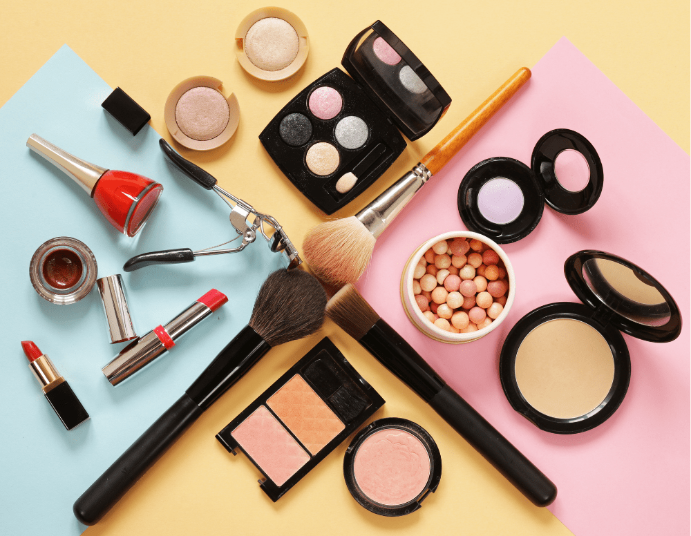 Assorted makeup products including lipsticks, nail polish, eyeshadow palettes, brushes, compact powders, and blushes arranged on a pastel background.