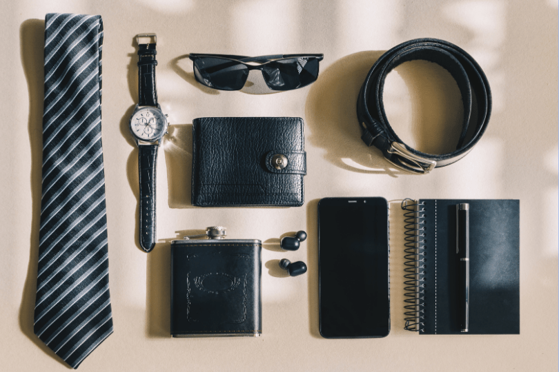 Flat lay of men's accessories including striped necktie, wristwatch, sunglasses, leather wallet, belt, flask, wireless earbuds, smartphone, spiral notebook, and pen arranged on a neutral surface