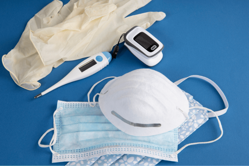 Personal protective equipment including N95 mask, surgical mask, patterned face mask, disposable gloves, digital thermometer, and pulse oximeter on a blue background.