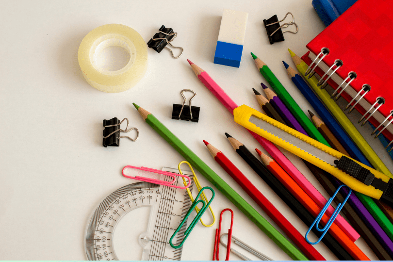 Assorted stationery items including colored pencils, binder clips, eraser, spiral notebook, utility knife, ruler, protractor, and paper clips arranged on a white surface