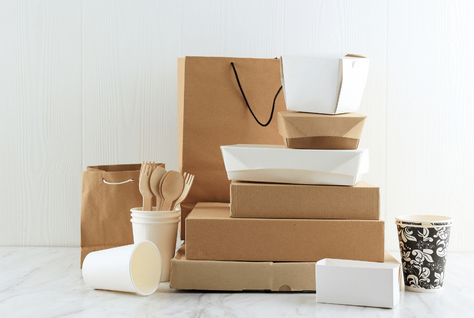 Eco-friendly disposable packaging including cardboard boxes, paper cups, food containers, and wooden cutlery arranged on a white wooden surface.