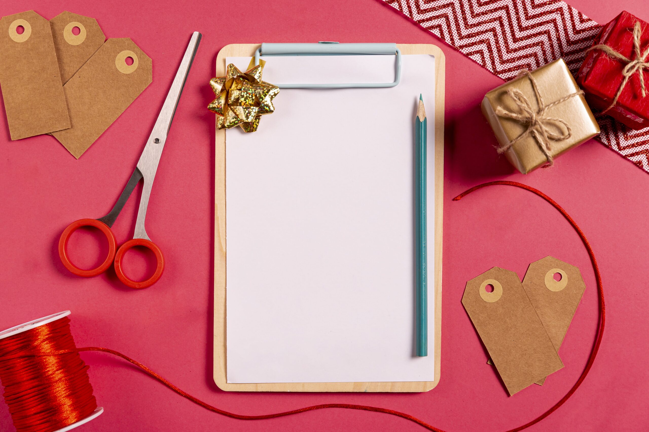 Flat lay of gift wrapping supplies including scissors, ribbon, gift tags, wrapped presents, and a clipboard with paper on a pink background.