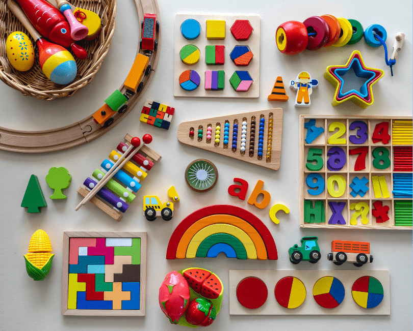 Assorted educational toys including wooden puzzles, abacus, xylophone, geometric shapes, toy vehicles, alphabet letters, and colorful fruit-shaped pieces arranged on a flat surface
